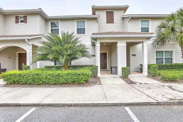 A multi-story residential building with a central entrance, a covered porch, and a small garden area with a palm tree. - Let's Glo Now Adventure Cove Villa 4 Bedroom Townhouse (Kissimmee)