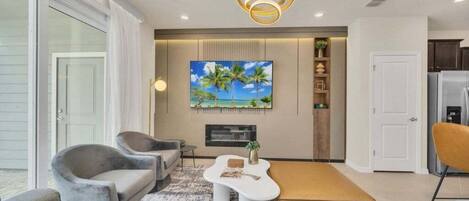 Una sala de estar moderna con un televisor grande, asientos cómodos y una cocina visible a través de una puerta abierta.