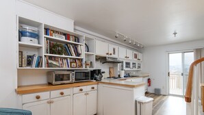 A modern kitchen with white cabinetry, a wooden countertop, and a built-in microwave. There is a bookshelf filled with books and decorative items, and a small trash bin is visible. - 6 Village Edge 2 Bedroom Condo (Warren)