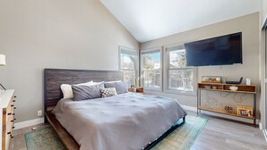 A bedroom with a large bed, a TV mounted on the wall, and a window with a view of the outside. - Snowcreek V #888 3 Bedroom Condo (Mammoth Lakes)
