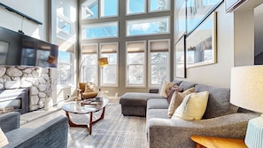 A spacious living room with a stone wall, large windows, a flat-screen TV, and a comfortable seating area. - Snowcreek V #888 3 Bedroom Condo (Mammoth Lakes)