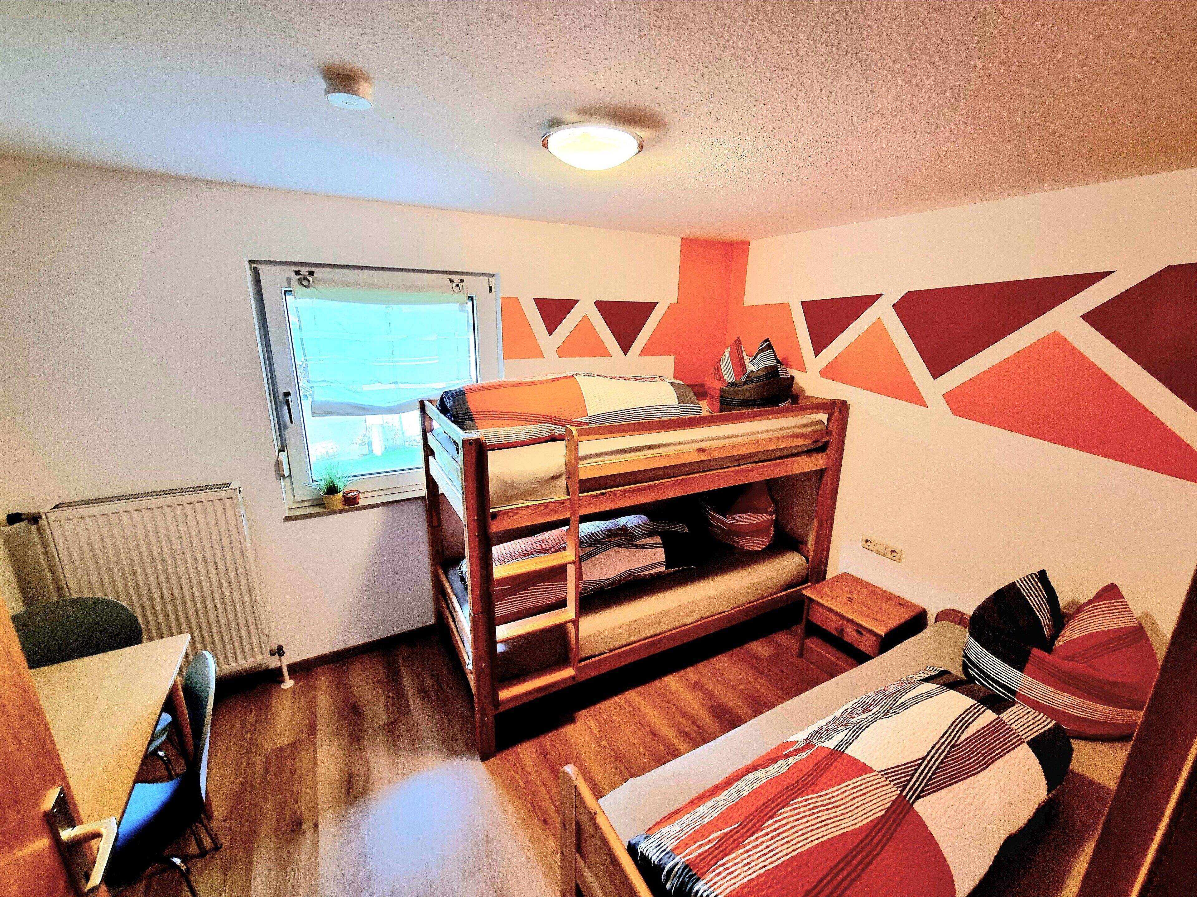 A bunk bed with a desk and chair, a radiator, a window, and geometric wall decorations.