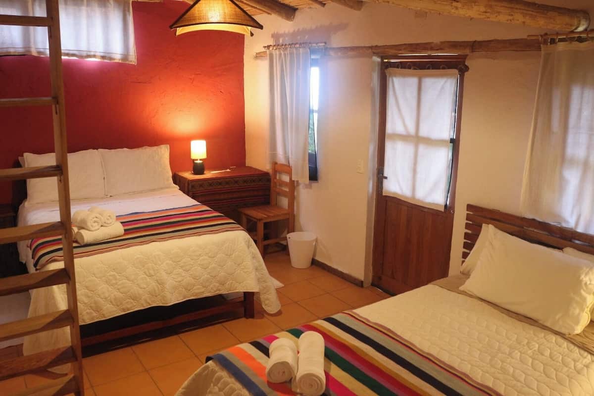 A room with two beds, a wooden ladder, a small table with a lamp, and a window with curtains.