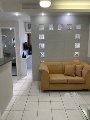 Een moderne woonkamer met een beige bank, een betegelde vloer, een grijze textuurwand met decoratieve glazen panelen, en een zichtbare keuken met een raam en een haardroger.