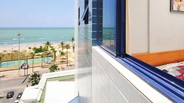 A room with a large window overlooking a beach, a bed, and a painting on the wall. - Flat Com Vista Mar No Everest (Recife)