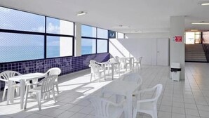 A spacious room with large windows, white plastic chairs, and tables arranged for dining. - Flat Com Vista Mar No Everest (Recife)