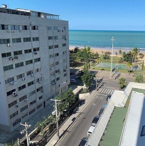 A multi-story building with air conditioning units, a street with cars, a pedestrian crossing, and a beach with palm trees in the background. - Flat Com Vista Mar No Everest (Recife)