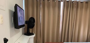 A room with a mounted TV, a standing fan, and a curtain. - Flat Com Vista Mar No Everest (Recife)