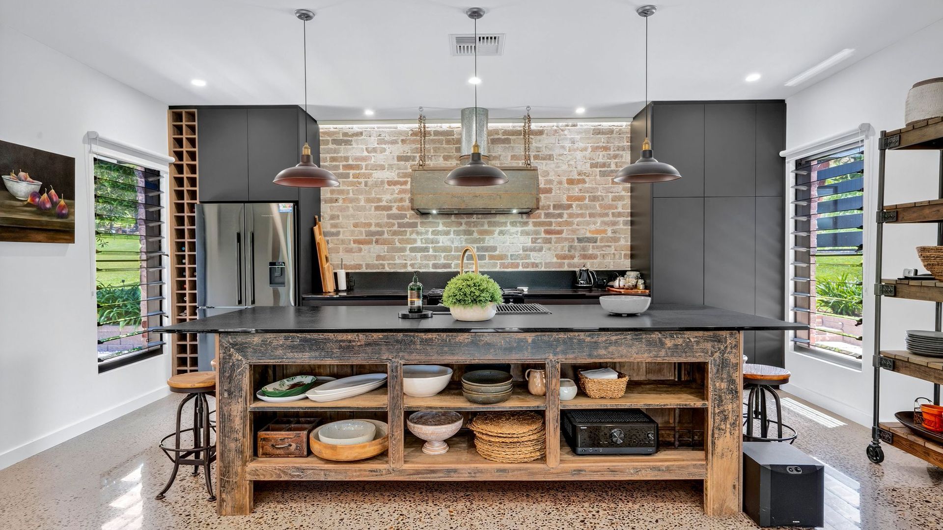 Cozinha com ilha central, luminárias pendentes, parede de tijolos e vista para o exterior.