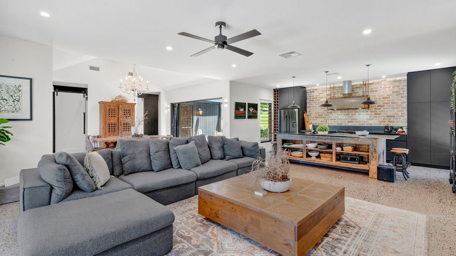 A modern living room with a sectional sofa, a wooden coffee table, and a kitchen area with a brick wall and bar stools.
