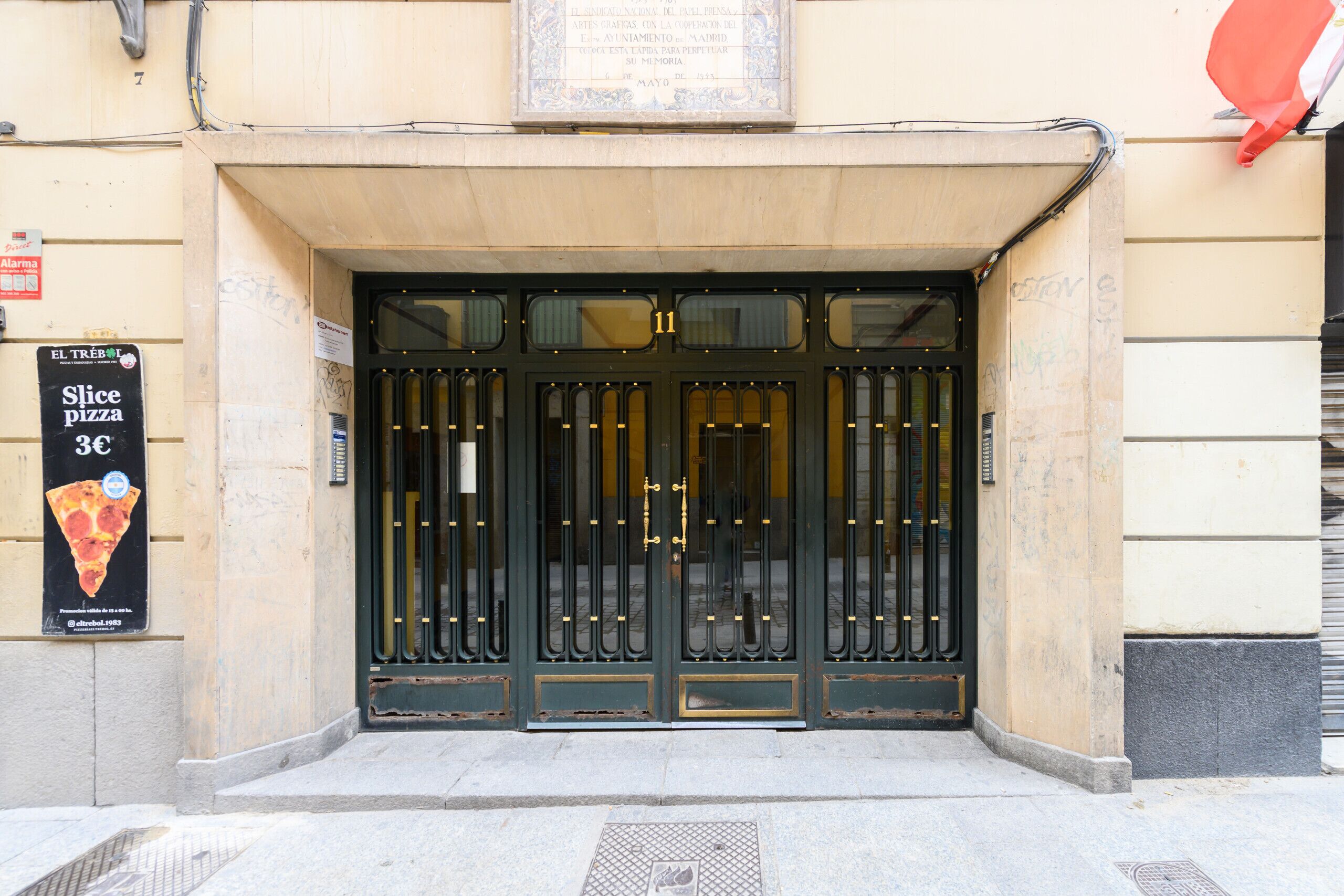 Entrada de un edificio con un gran portón negro, un letrero que anuncia pizza al corte y una placa sobre la puerta.