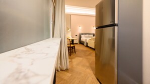 Ein kompaktes Hotelzimmer mit Bett, Kühlschrank, Esstisch mit Stühlen und einer kleinen Sitzecke mit Sofa und Lampe.