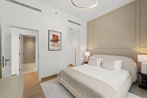 Quarto de hotel moderno com uma cama grande, uma mesa de cabeceira com luminária, uma obra de arte emoldurada na parede e uma porta que dá para outro cômodo.