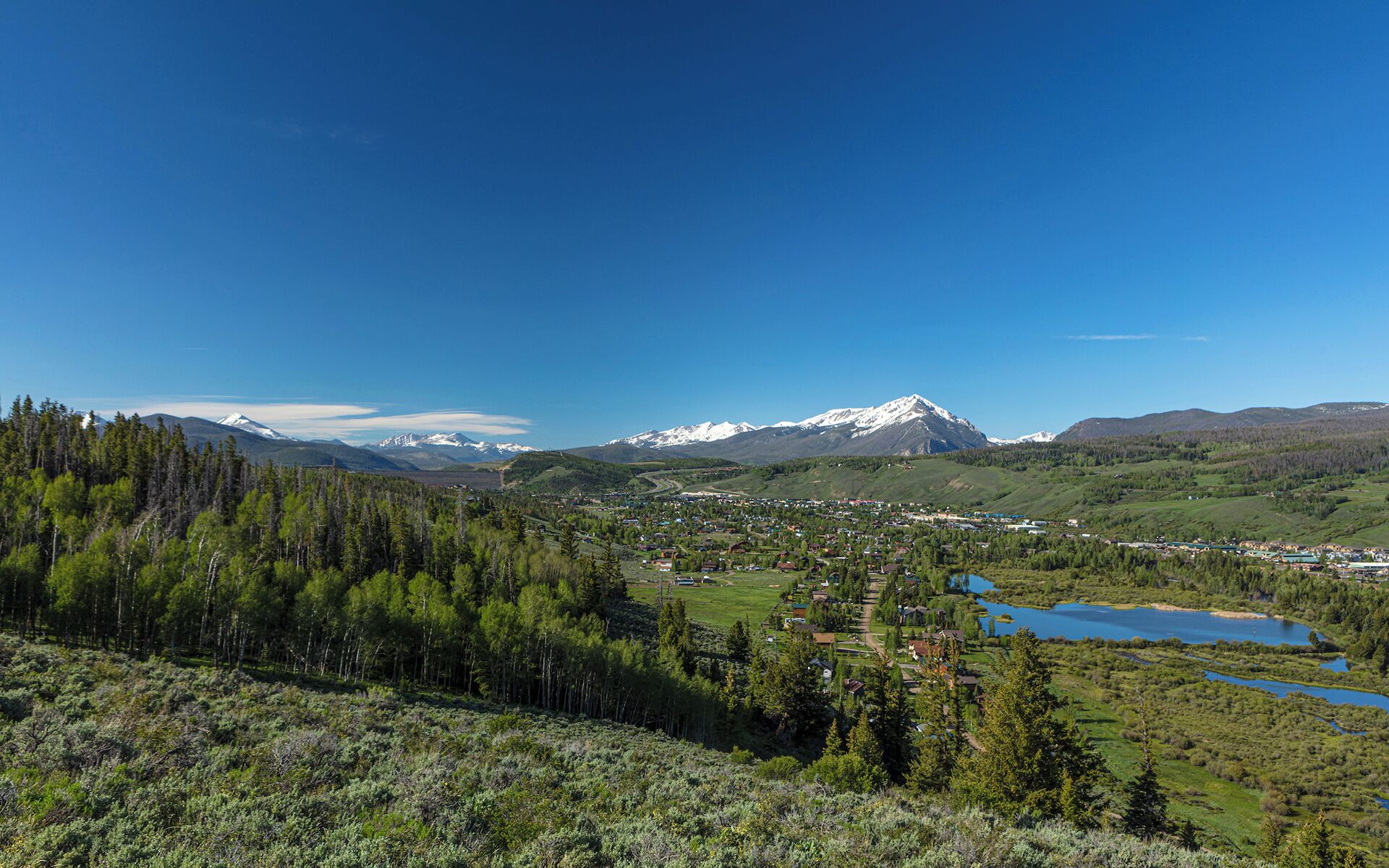 A mountainous landscape with a town in the valley, a lake, and snow-capped peaks in the distance.