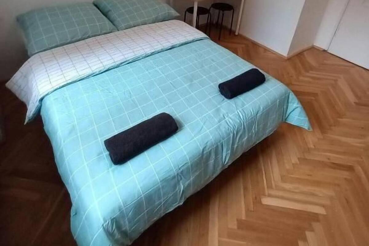 Un llit individual amb un edredó blau i dos coixins, un petit escriptori de fusta amb una cadira, i una tetera sobre l'escriptori.