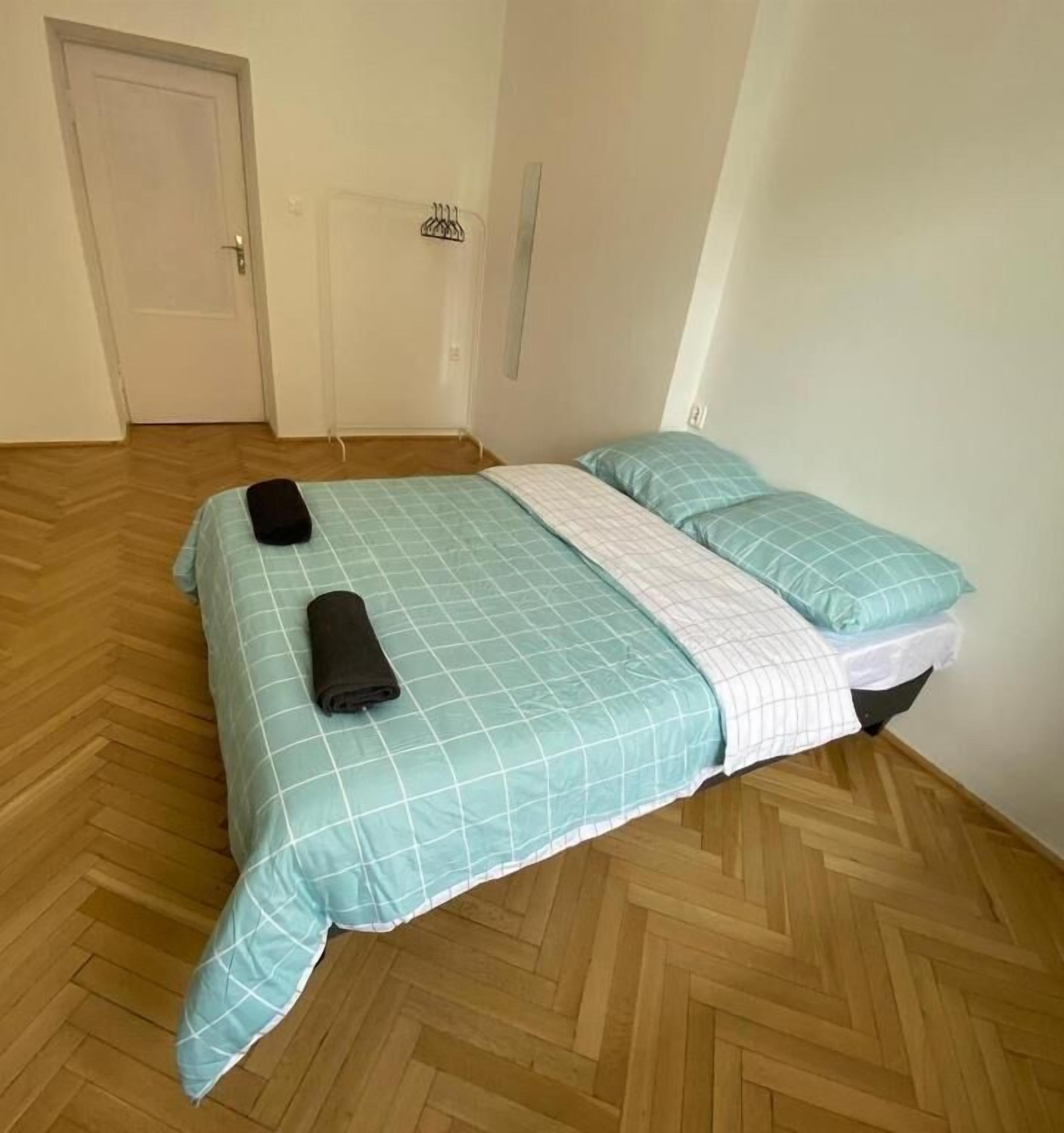 Een kleine, verzorgde slaapkamer met een houten visgraatvloer, een bed met een blauw-witte geruite spreidek en twee opgerolde handdoeken op het bed.