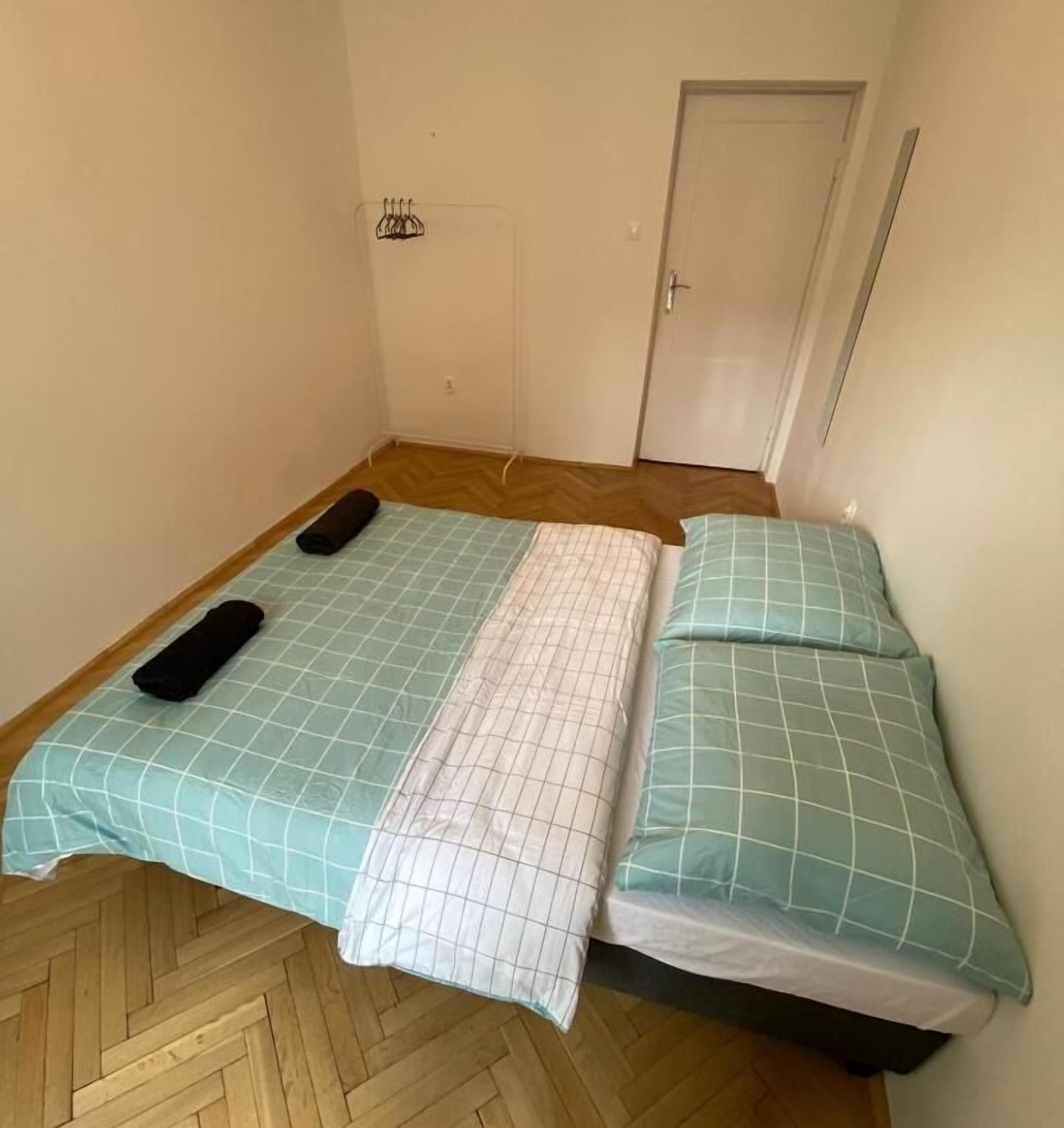 Habitació petita amb un llit individual, un sòl de fusta en rellevat, una porta blanca i un penjador amb ganyots.