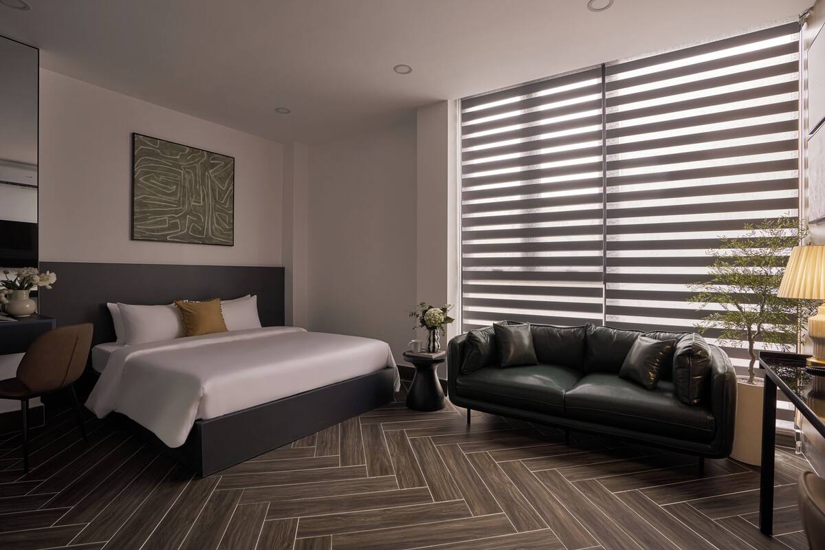 A modern hotel room with a large bed, a dark leather sofa, a small side table with a vase, and a large window with blinds.