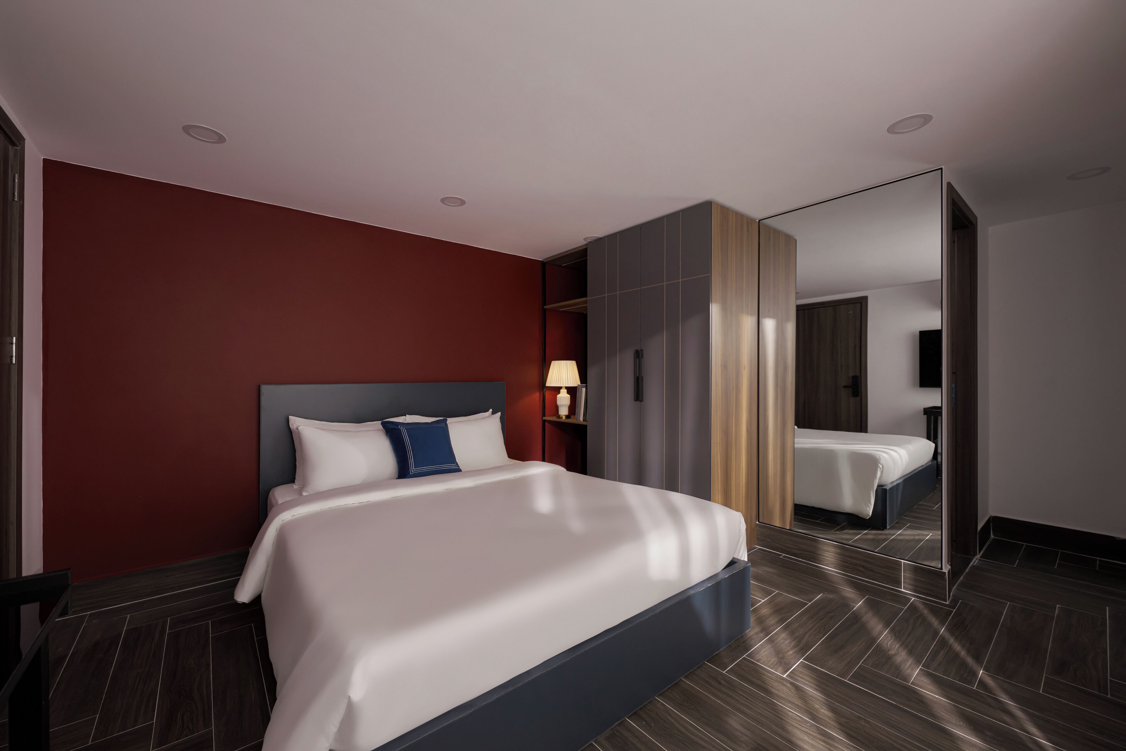 A modern hotel room with a large bed, a nightstand with a lamp, a wardrobe, and a mirror.
