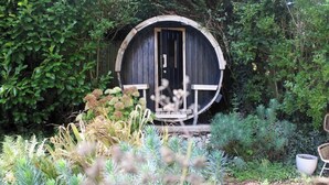 A rustic wooden structure with a circular door, surrounded by lush greenery and various plants. - Lewes Sauna House · Characterful Interiors, Castle Views (East Sussex)