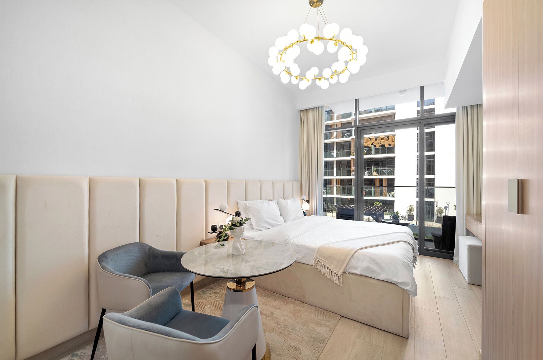 Una habitación de hotel moderna con una cama grande, una mesa redonda, dos sillas, una araña y vistas de la ciudad a través de amplias ventanas.