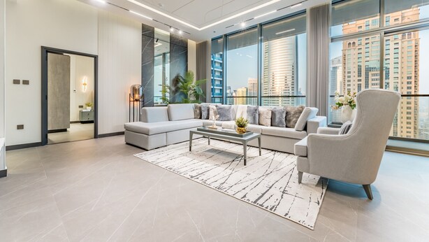 A modern living room with a sectional sofa, a coffee table, and a large window offering a city view. - Luxurious High-Floor 3BR with Assistant Room, Showcasing Lake & City Views (Dubai)