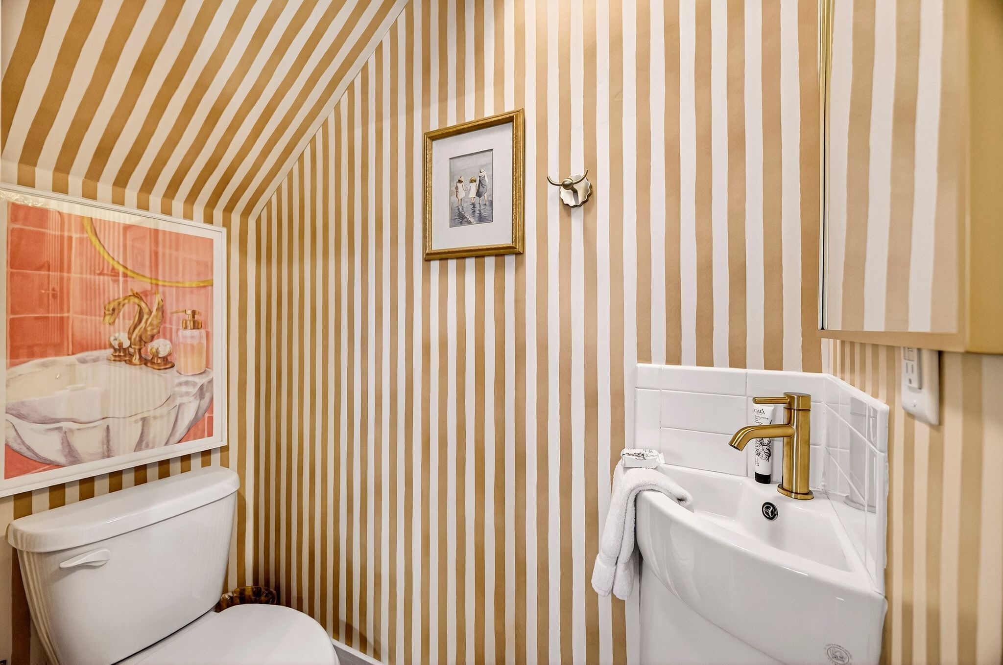 A bathroom with striped wallpaper, a framed picture, a toilet, a sink with a gold faucet, and a decorative shell on the wall.