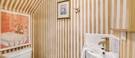 A bathroom with striped wallpaper, a framed picture, a toilet, a sink with a gold faucet, and a decorative shell on the wall.