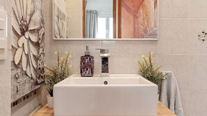 Ein Badezimmer mit einem rechteckigen Waschbecken, einem Spiegel darüber und einer dekorativen Wandverkleidung links.