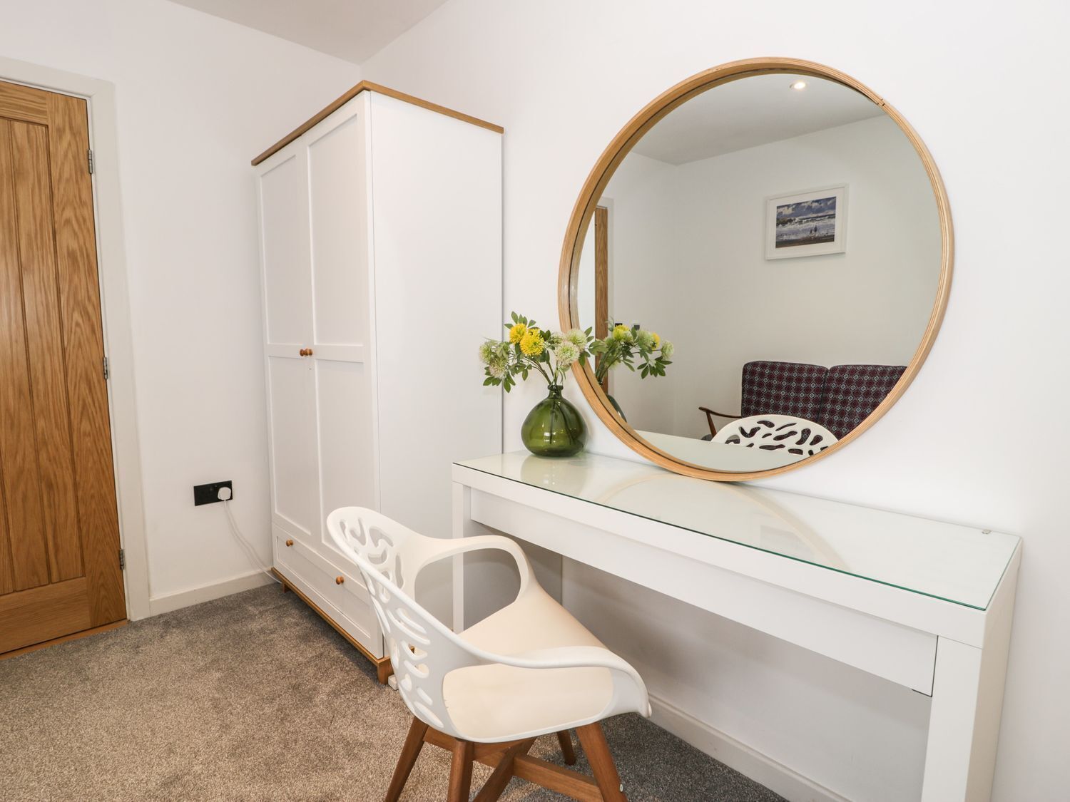 Un dormitorio con un armario blanco, un escritorio de vidrio, una silla, un espejo redondo y un florero con flores.