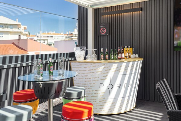 A rooftop bar with a bar counter, glasses, bottles, and a sign prohibiting smoking. - Porta 30 (Oeiras)