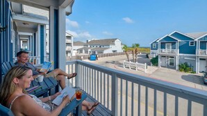 Een balkon met twee personen die lezen, een helder uitzicht op het strand en een rij blauwe gebouwen op de achtergrond.