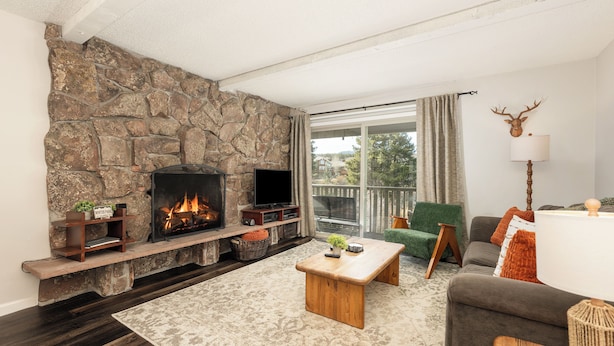 A cozy living room with a stone fireplace, a flat-screen TV, comfortable seating, and a view through a large window. - Hi Country Haus Unit 2110 Spacious 2 bed, river views walk to downtown (Winter Park)