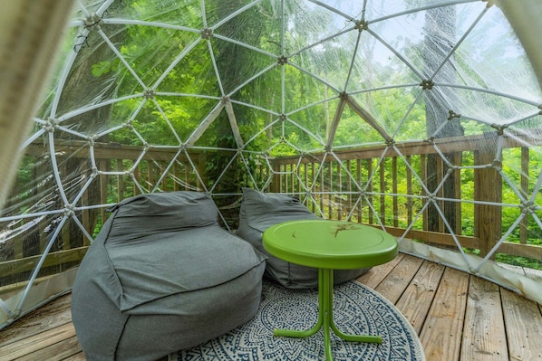 Een geodetische koepelstructuur met een houten terras, grijze zachte zitplaatsen en een groene tafel.