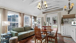 Ein gemütliches Wohnzimmer mit einem grünen Sofa, einem Holzespeisesaal und Stühlen. Große Fenster mit Vorhängen, eine Chandelier und ein kleiner Beistelltisch mit Lampe sind ebenfalls vorhanden.