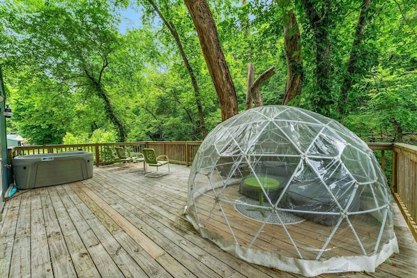 Estructura de cúpula geodésica transparente sobre una cubierta de madera, con un jacuzzi en su interior, rodeada de exuberantes árboles verdes.