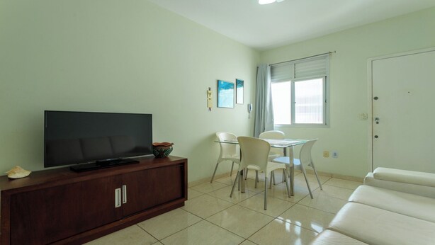 A living room with a flat-screen TV on a wooden cabinet, a dining table with chairs, a white sofa, and a window with blinds. - Apto Pet Friendly com Garagem na Praia do Morro (Guarapari)