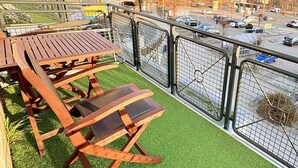 En balkong med ett träbord och stolar, omgiven av ett metallstaket, med utsikt över en parkeringsplats och byggnader.