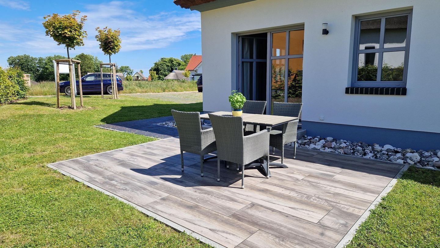 Een modern huis met een patio, een tafel en stoelen, een kleine tuin en een nabijgelegen geparkeerde auto.