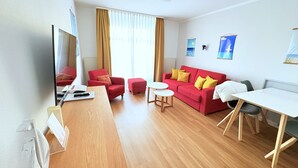 Ein modernes Wohnzimmer mit Holzboden, einem roten Sofa mit gelben Kissen, einem weißen Tisch und einem Fernseher links.