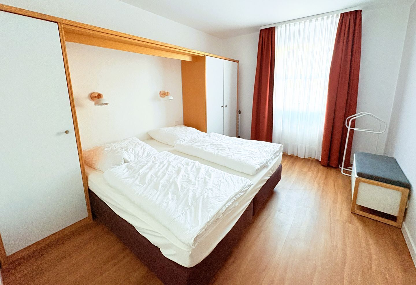 A double bed with white bedding in a room with wooden flooring and a window with red curtains.