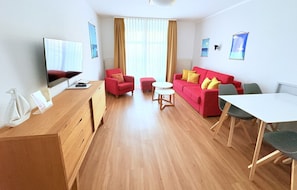 Un salon moderne avec un canapé rouge, un bureau en bois, un téléviseur et un coin repas comprenant une table et des chaises blanches.