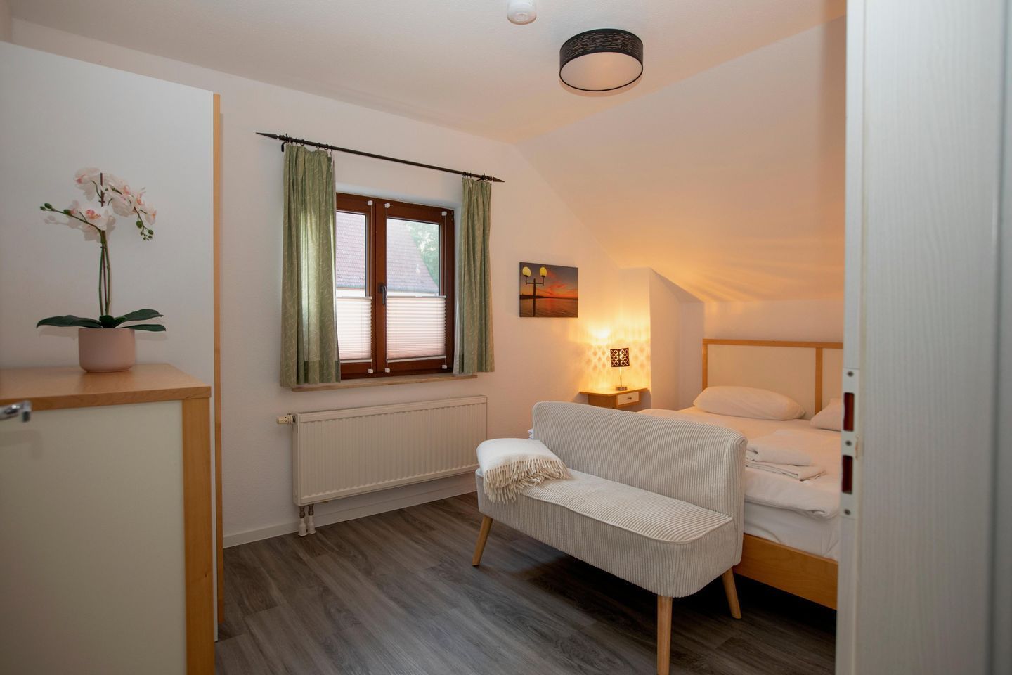 Ein Schlafzimmer mit einem Bett, einem Nachttisch, einer Lampe, einer Heizung, einem Fenster mit Vorhängen und einer Pflanze.