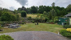 Ein Garten mit befestigter Fläche, Bänken und einem kleinen Gebäude.