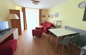 A compact hotel room with a desk, a red sofa, a dining table with chairs, a lamp, and a clock on the wall. - Apartment 12 in Haus Hühnergott - top location for families - Holiday apartment 12 - (Sellin)