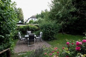 Eine Terrasse mit Tisch und Stühlen, umgeben von üppigem Grün und blühenden Pflanzen.