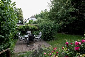A patio area with a table and chairs, surrounded by lush greenery and flowering plants. - Milana Apartment - Perfect for Two, Parking - Milana Apartment - Sun, (Putbus)