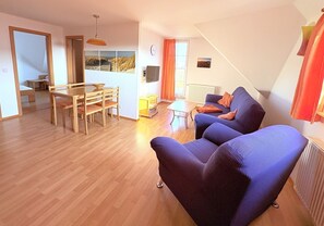 Ein Wohnzimmer mit einer Couch, einem Esstisch mit Stühlen, einem kleinen Tisch und einem Fenster mit Vorhängen.