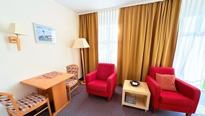 Hotellihuone, jossa on puinen työpöytä, kaksi punaista nojatuolia, pieni pöytä lampun kanssa ja suuri ikkuna, jossa on verhot.