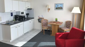 Kompakti keittiö, jossa on valkoiset kaapit, mikroaaltouuni ja kahvinkeitin. Siellä on pieni ruokailutila, jossa on kaksi tuolia ja pöytä, ja etualalla näkyy punainen tuoli.
